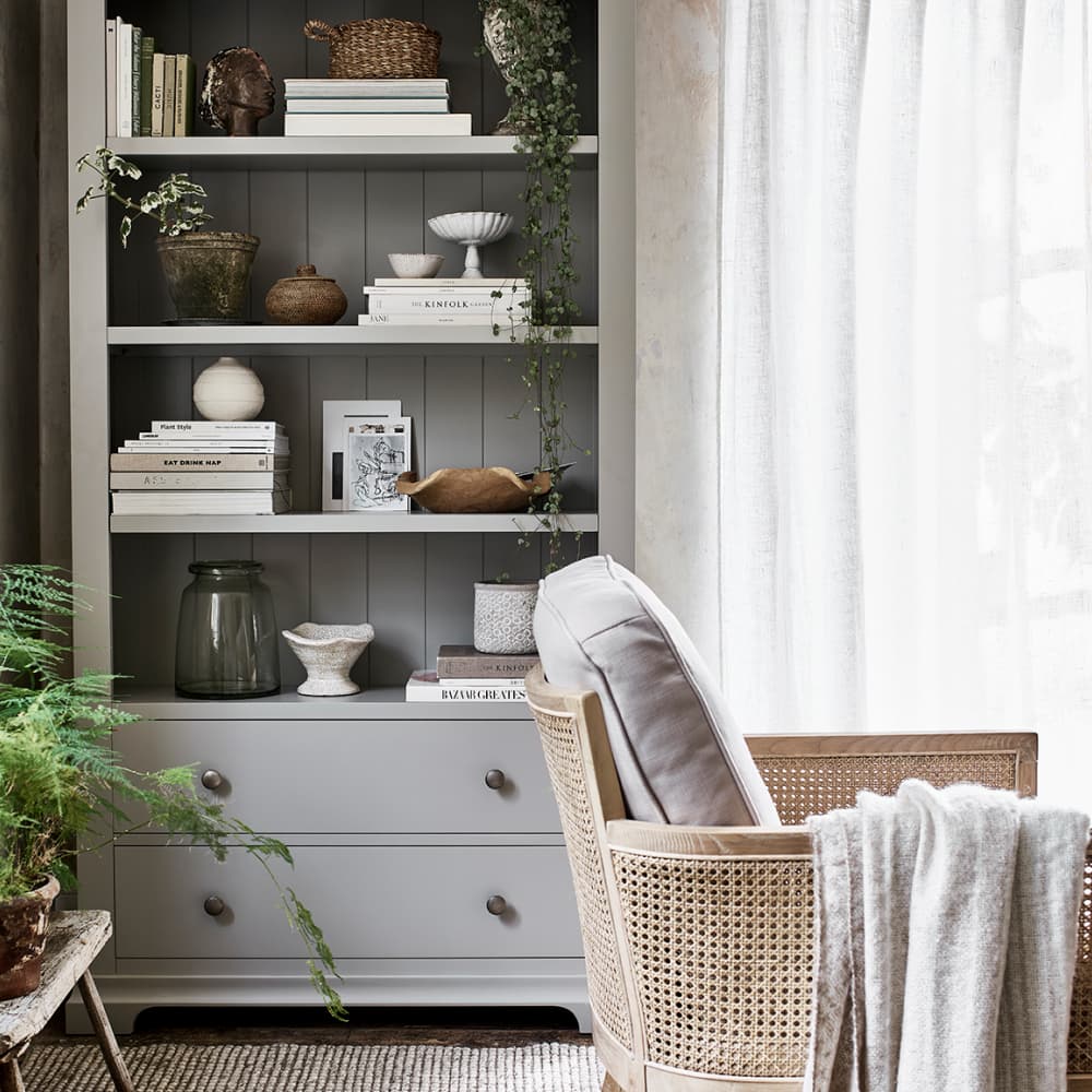 Cosy living room with a cushioned wicker chair, grey shelves displaying books, plants, and decor, and light curtains beside a window.