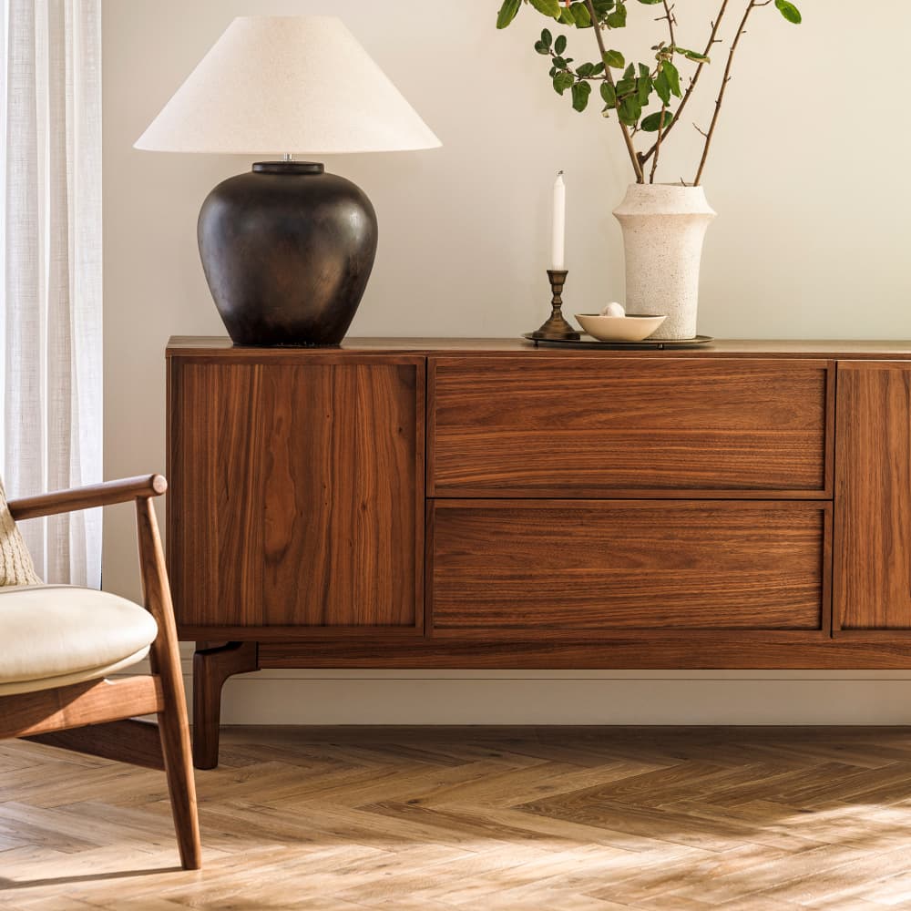 A dark wooden sideboard with a black lamp, a candle in a brass holder, and a tall vase with branches, next to a wooden chair on a parquet floor.