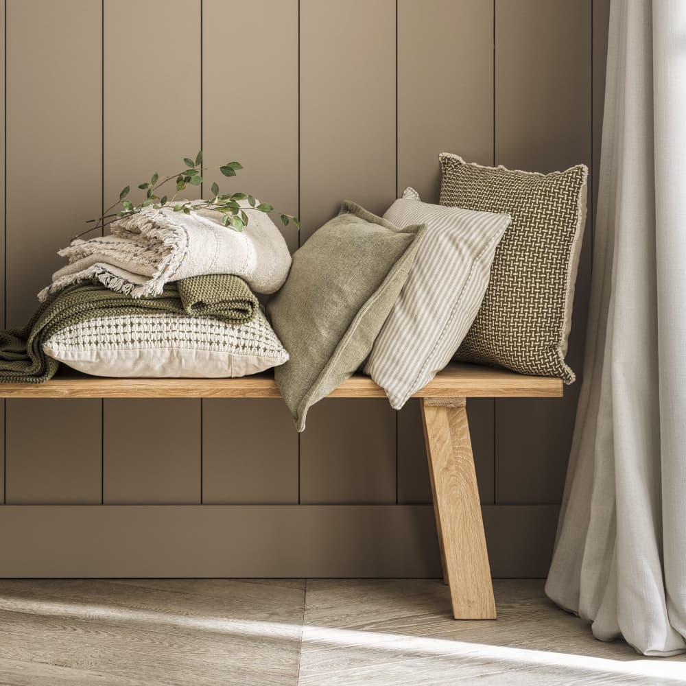 Wooden bench with assorted pillows and blankets in neutral tones, placed against a taupe panelled wall and a curtain beside.