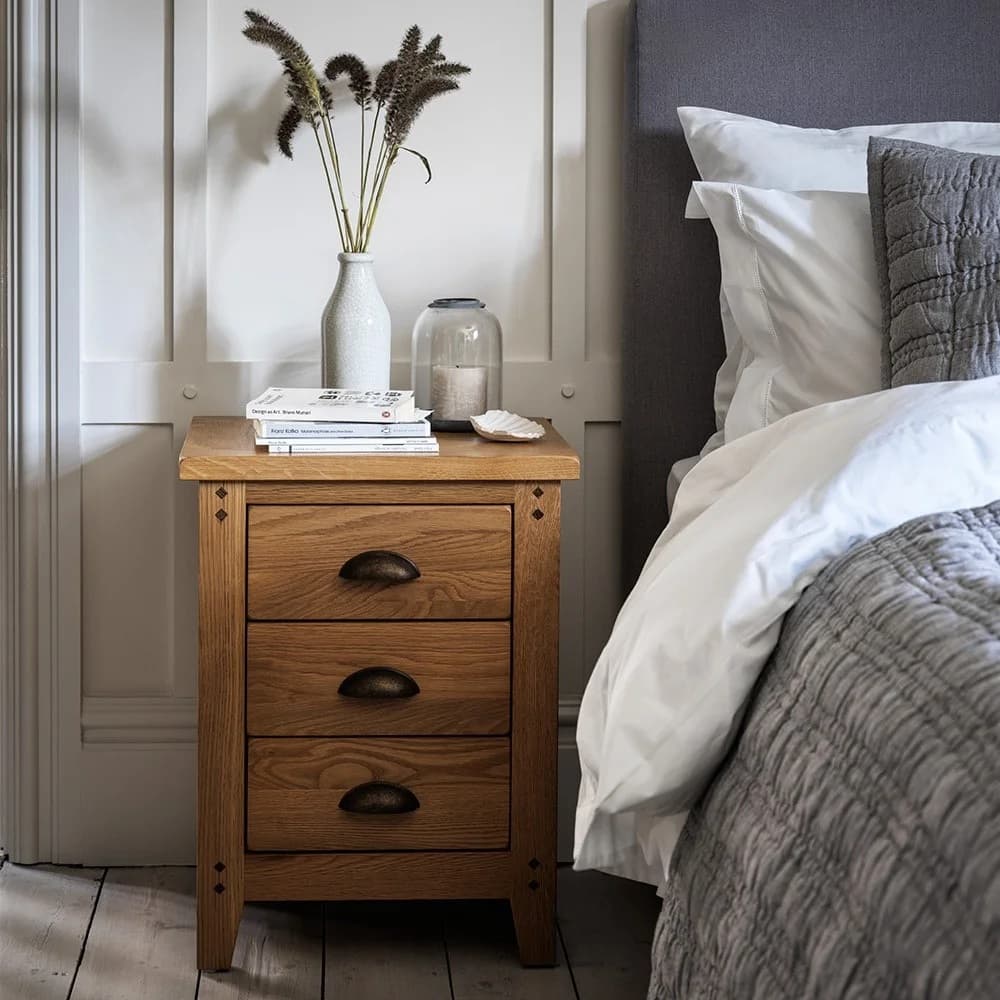 Wooden bedside table with three drawers, a vase with tall grass, books, and a candle. Next to a bed with white and grey bedding.