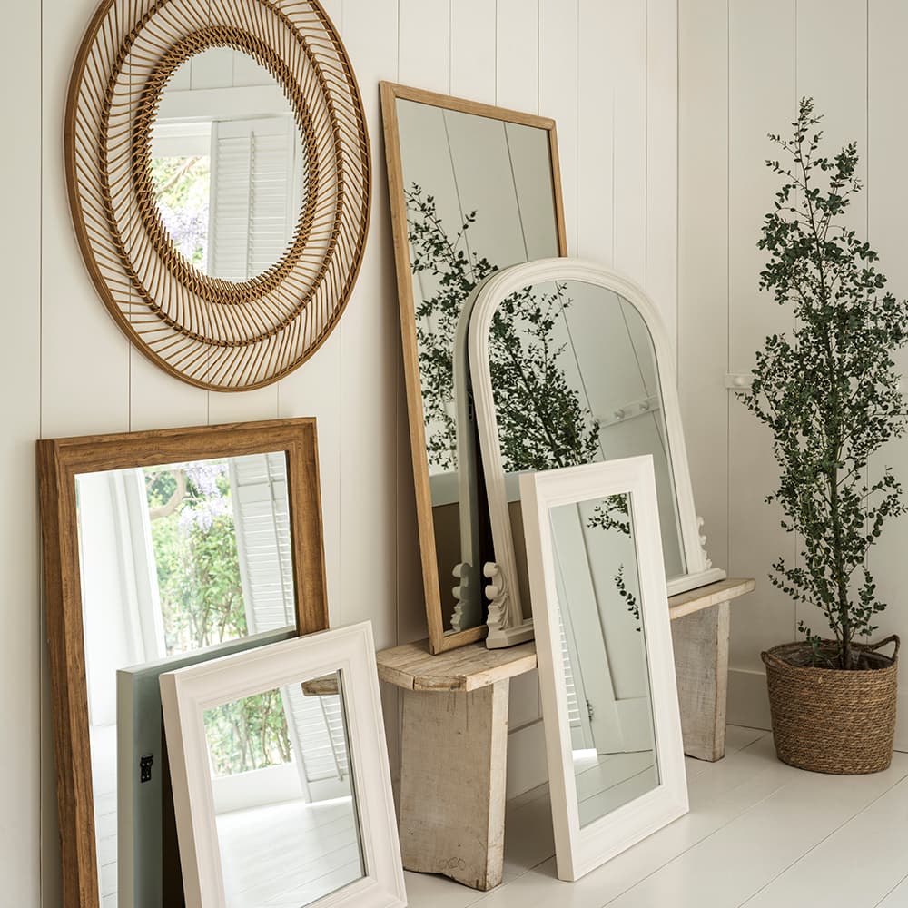 A collection of variously shaped mirrors leaning against a white panelled wall, next to a potted plant.