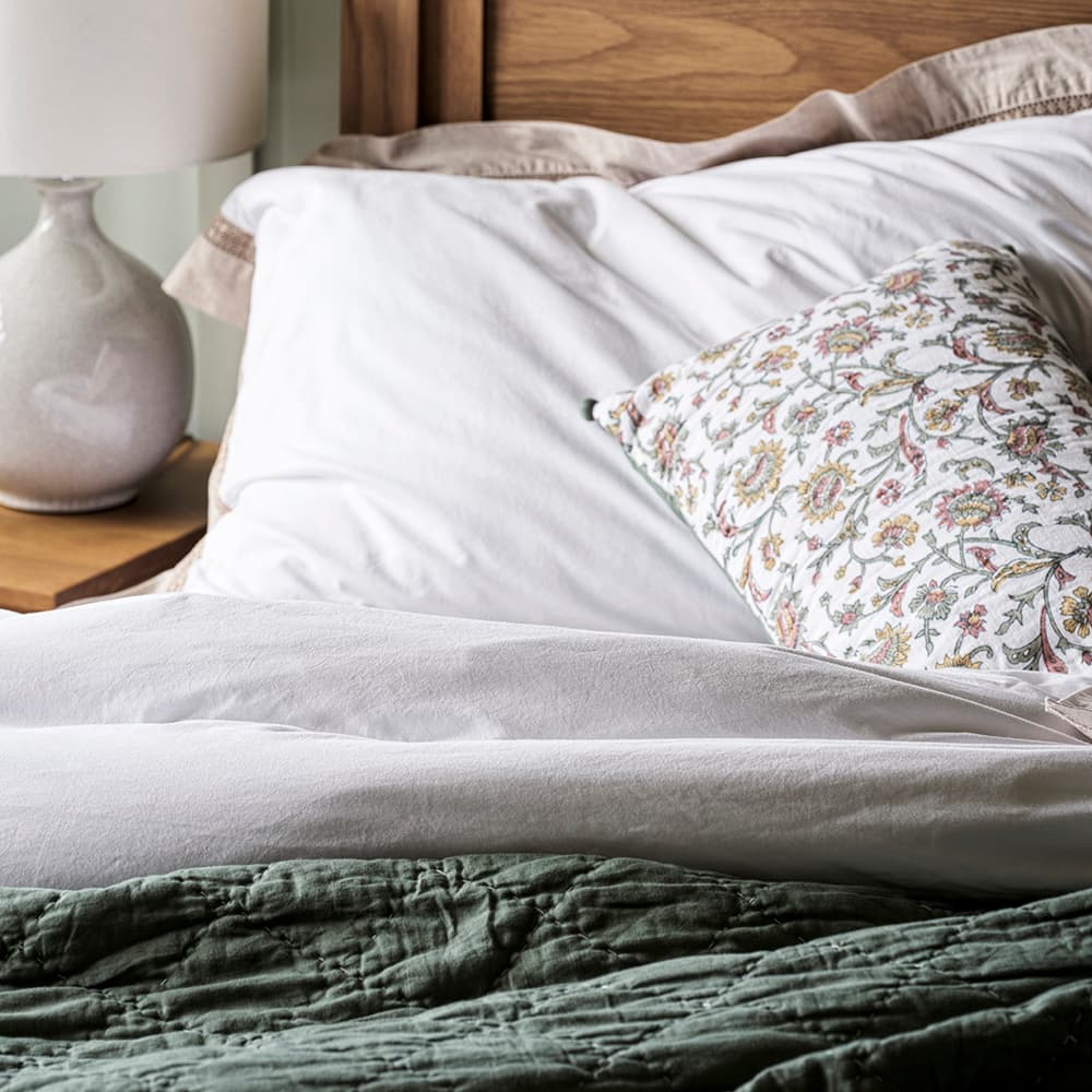 A neatly made bed with white sheets, a floral pillow, and a green quilt. A wooden headboard and a lamp on a nightstand are visible.