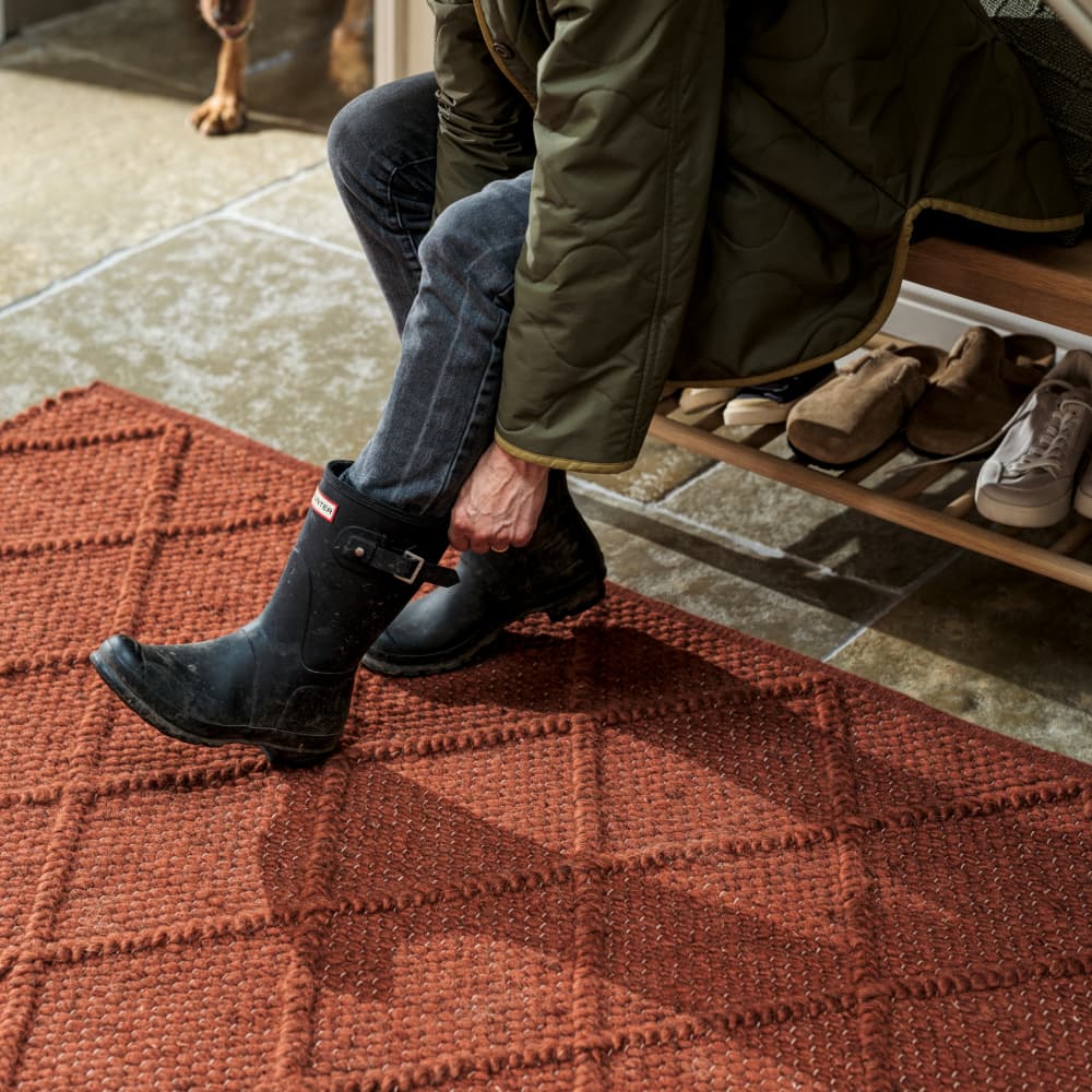 Person in a green coat puts on black boots while sitting on a bench near a red patterned rug, with shoes neatly arranged below.