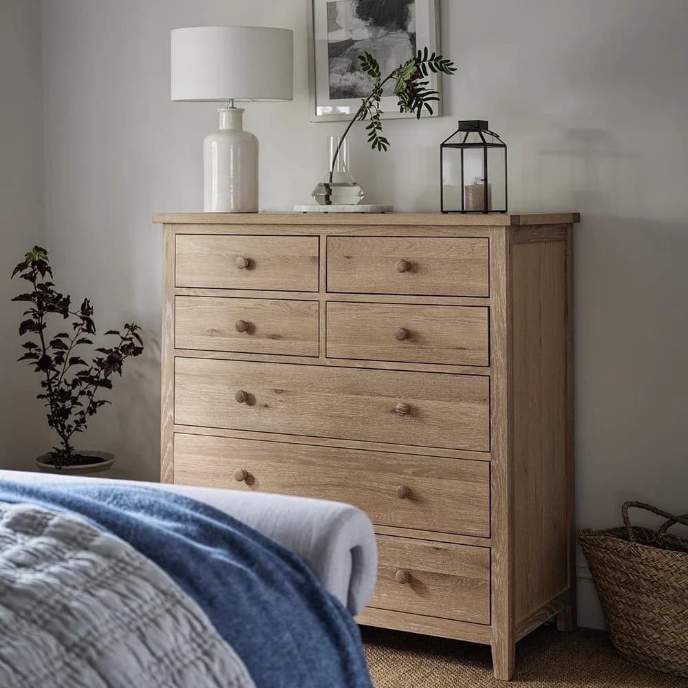 Wooden dresser with seven drawers, topped with a lamp, plant, and lantern. A bed with blue and white linens is visible in the foreground.