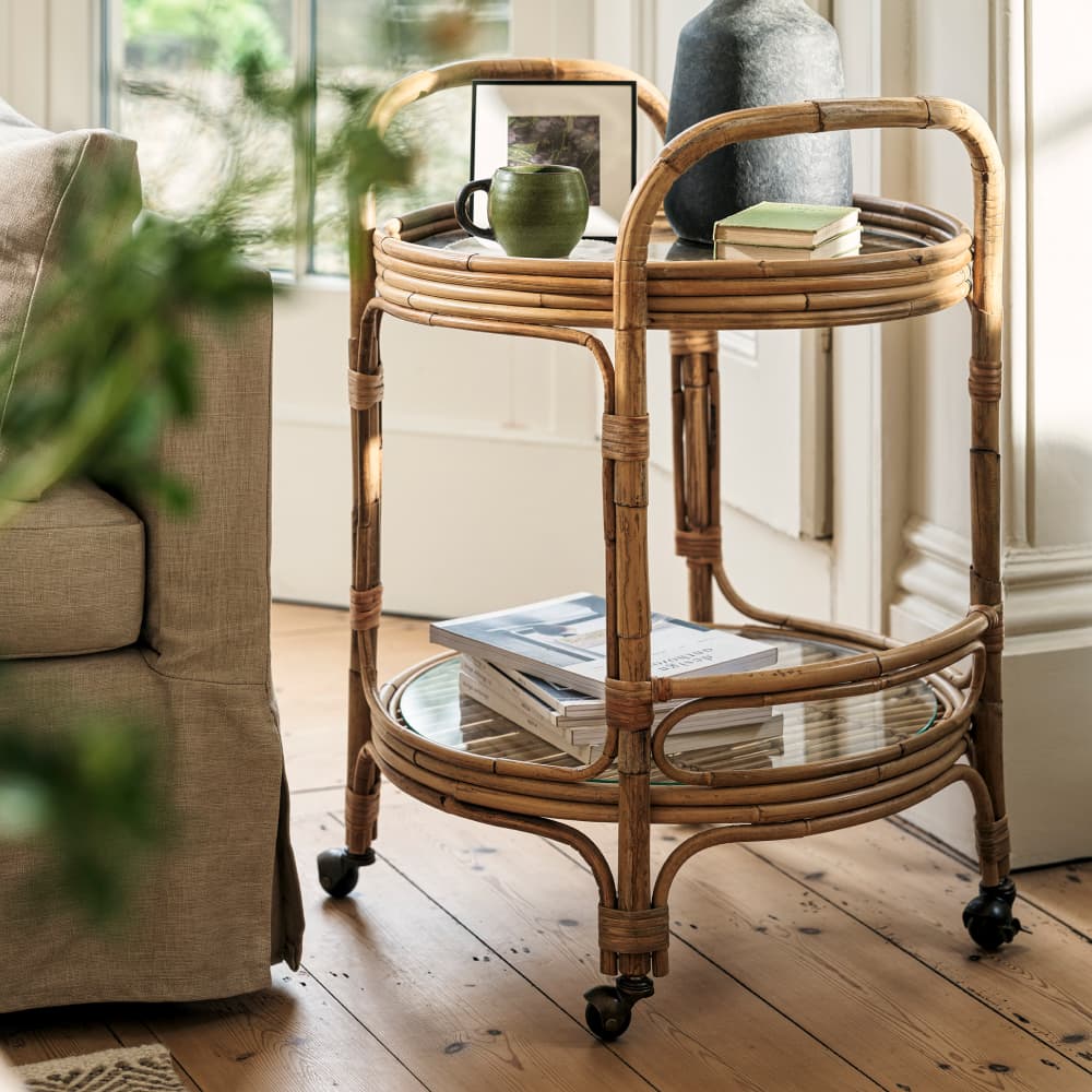 A side table cart with two shelves on wheels, holding a green mug, a dark vase, books, and a picture frame, beside a beige sofa in a sunlit room.