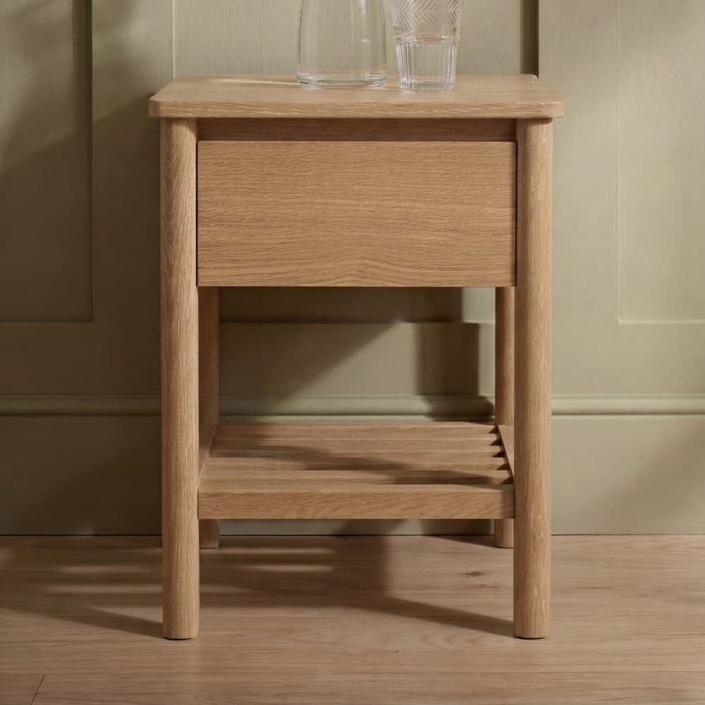 Wooden side table with a single drawer and a lower slatted shelf. Two clear glasses sit on top. The background is a light green wall.