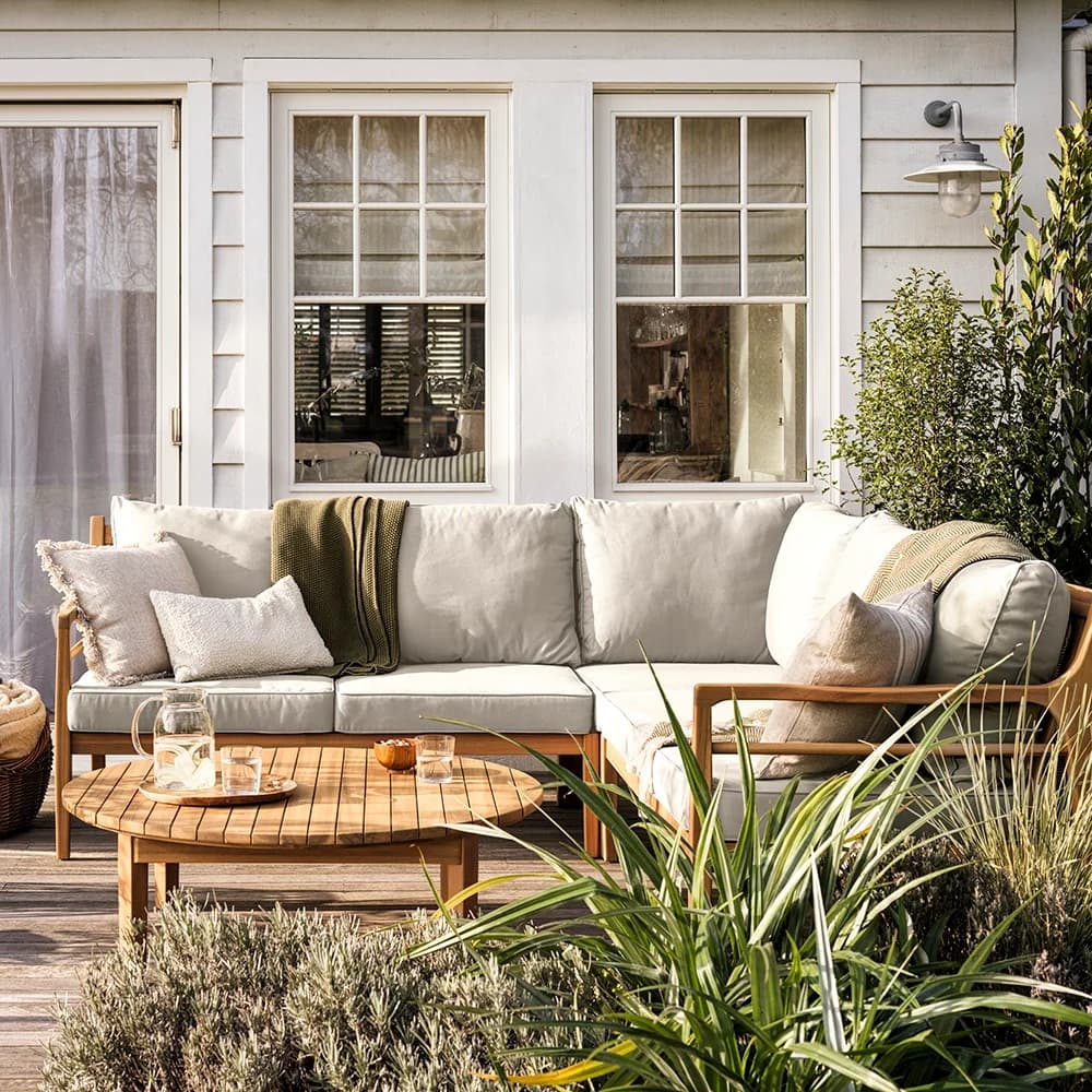 Outdoor patio with a beige sectional sofa, green and beige pillows, and a wooden coffee table, set against a house with large windows and greenery.