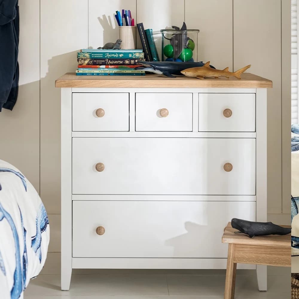 White dresser with wooden knobs, books, toy sharks, and a jar of colorful balls on top, next to a bed and wooden bench.