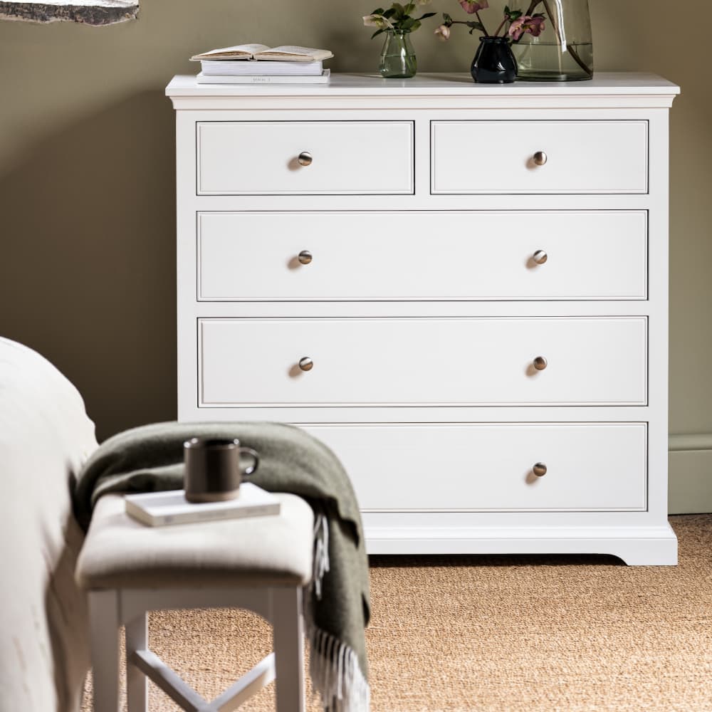 A white wooden dresser with five drawers, next to a bench with a mug and blanket. Carpeted floor and soft green walls in the background.