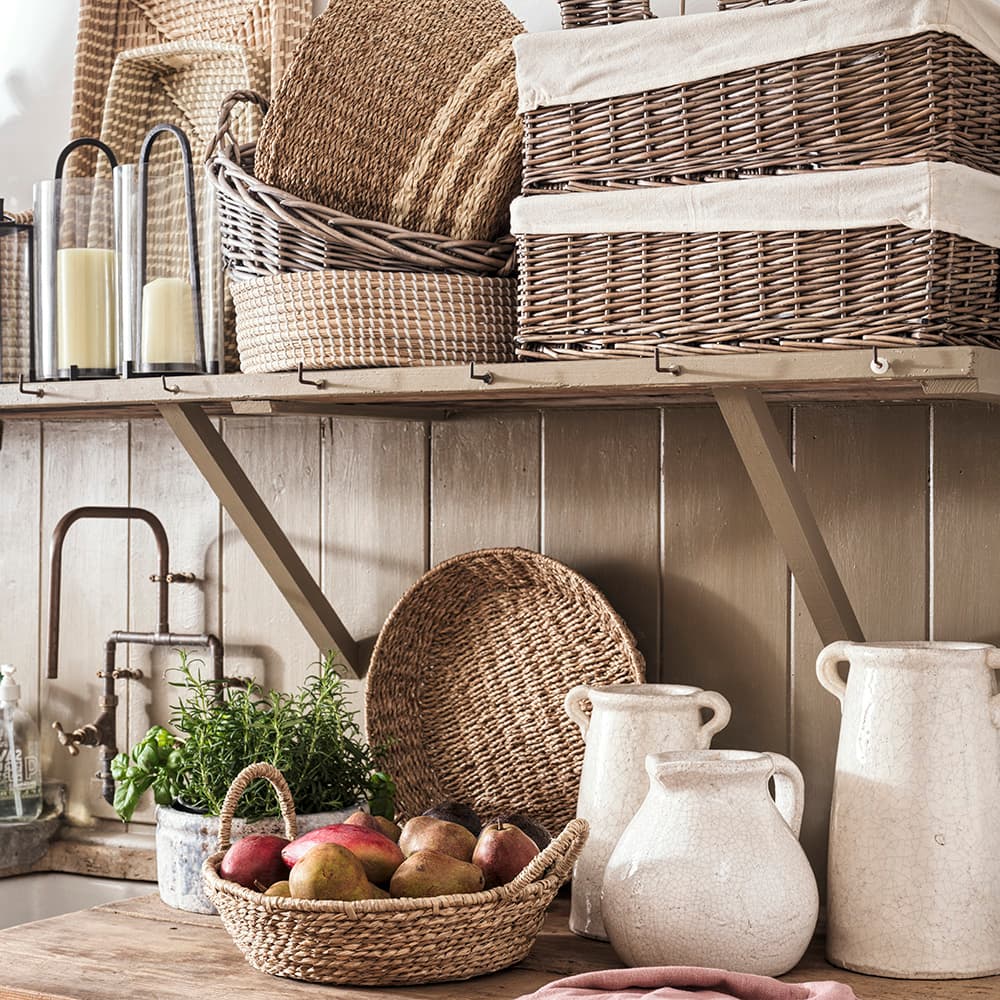 Rustic kitchen with woven baskets, candles, and pottery on wooden shelves. A basket of mangoes and a potted plant sit on the wooden countertop.
