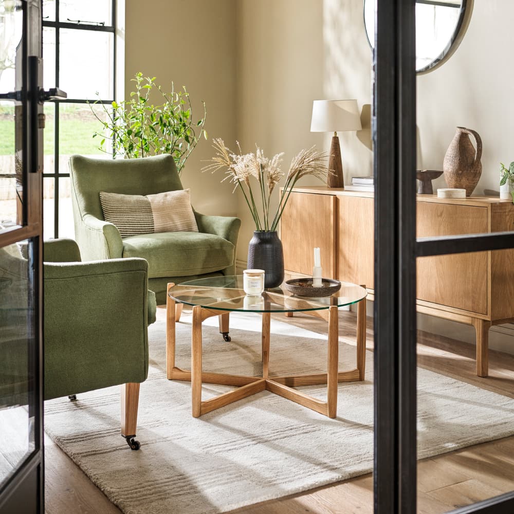 A cosy living room with green armchairs, a wooden coffee table with glass top, a wooden sideboard, and decorative plants, bathed in natural light.