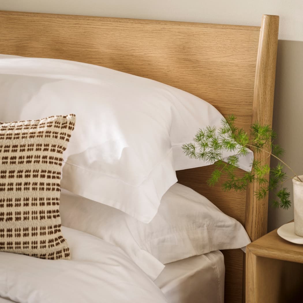 Close-up of a bed with white pillows, a patterned cushion, wooden headboard, and a small plant on a bedside table.