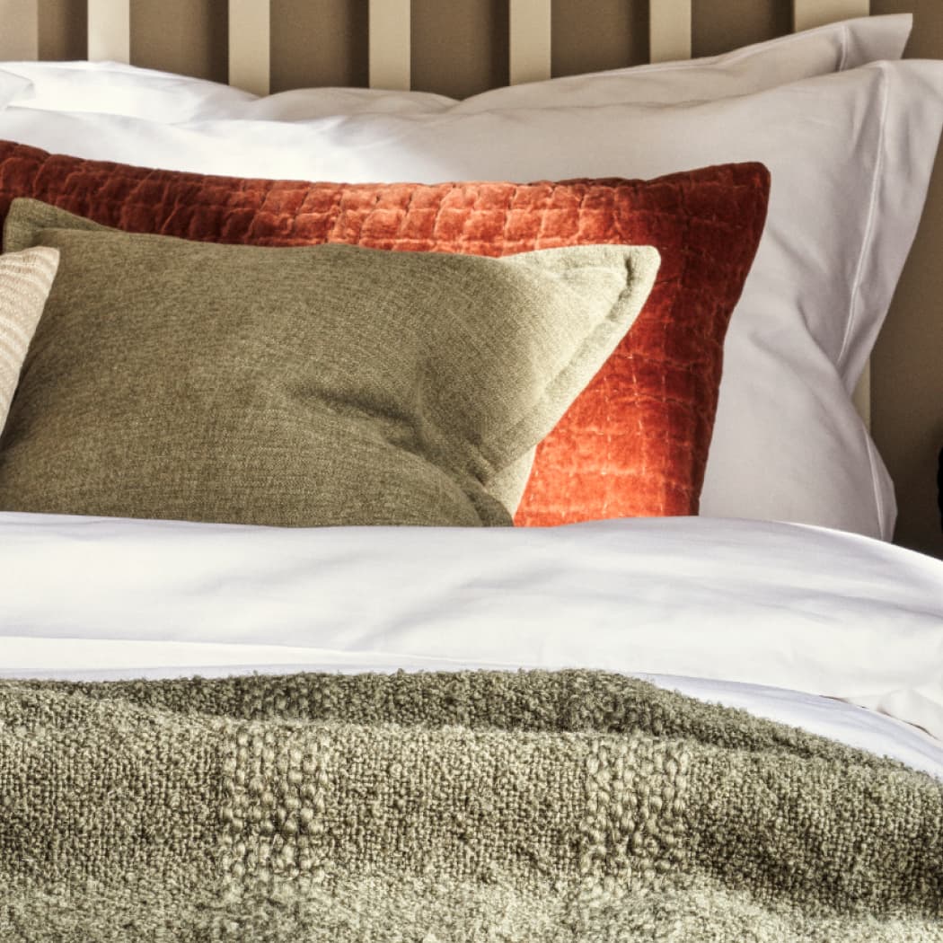 A neatly made bed with a red textured pillow, green pillow, white sheets, and a knitted green blanket.