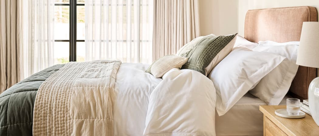 Cozy bedroom with a neatly made bed, layered pillows and blankets in neutral tones. A window with sheer curtains allows soft light to fill the room.
