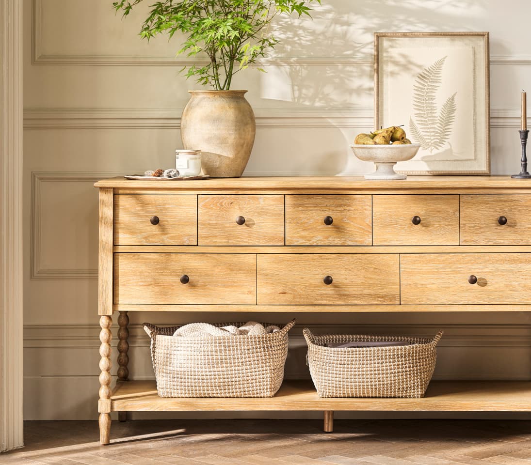Wooden sideboard with drawers, featuring a vase with greenery, framed fern print, bowl of pears, baskets underneath, and decorative items on top.