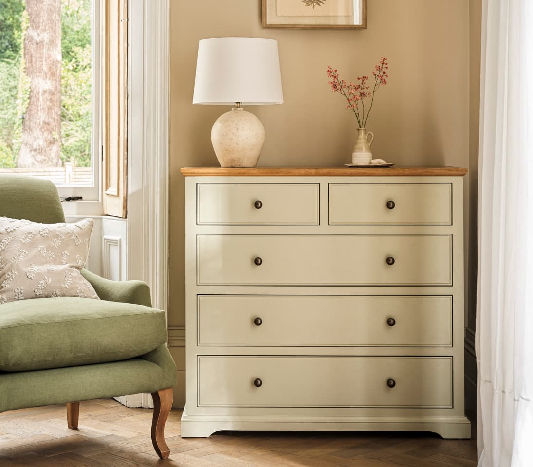 A beige dresser with a wooden top, featuring a lamp and a small vase with flowers, beside a green armchair with a patterned pillow.
