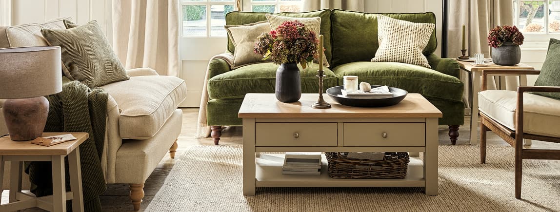 Cozy living room with green and beige sofas, wooden coffee table, decorative vases with flowers, and a soft rug. Natural light from large windows.