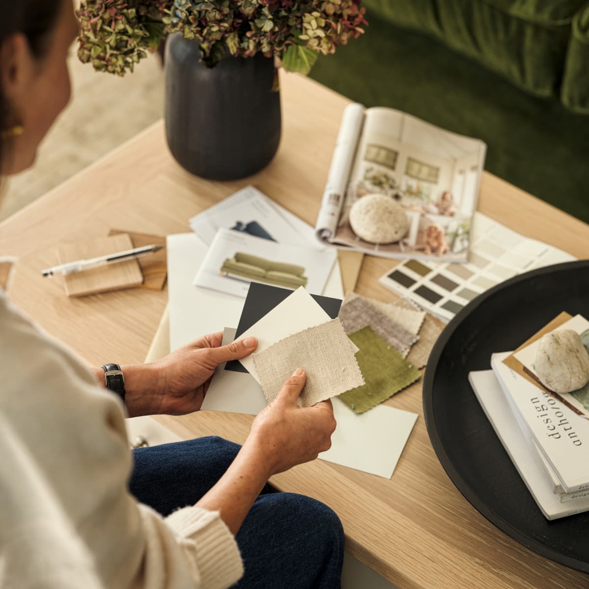 A person holding fabric swatches and color samples, sitting at a table with design magazines, notebooks, and a vase of flowers.