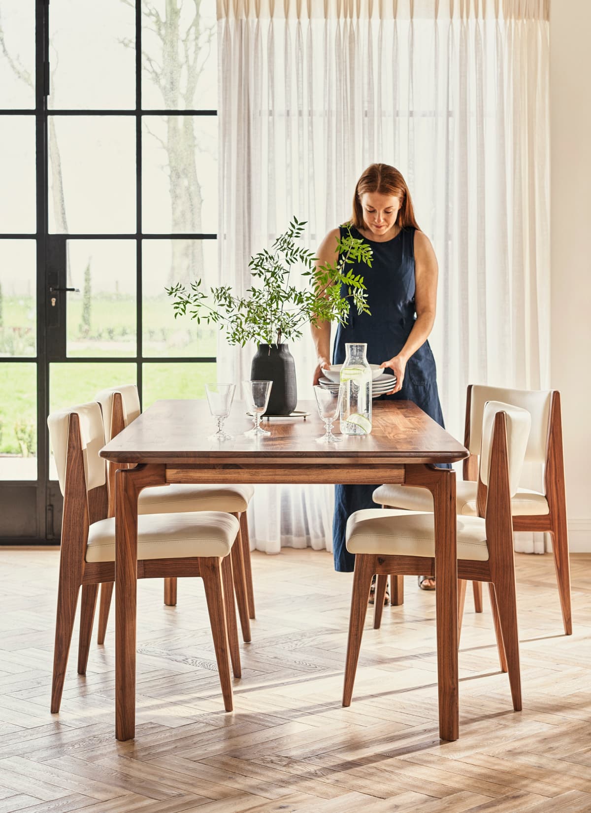 A woman sets a wooden dining table with plates and glasses, next to a vase with green foliage near large windows with sheer curtains.