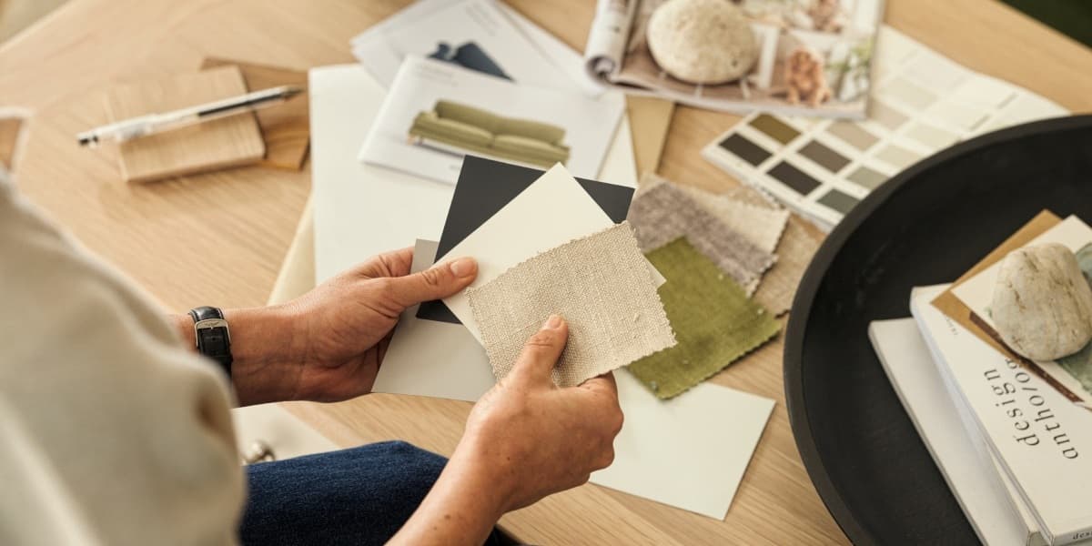 Person holding fabric and color swatches at a table with design magazines, samples, and a pen for interior decoration planning.