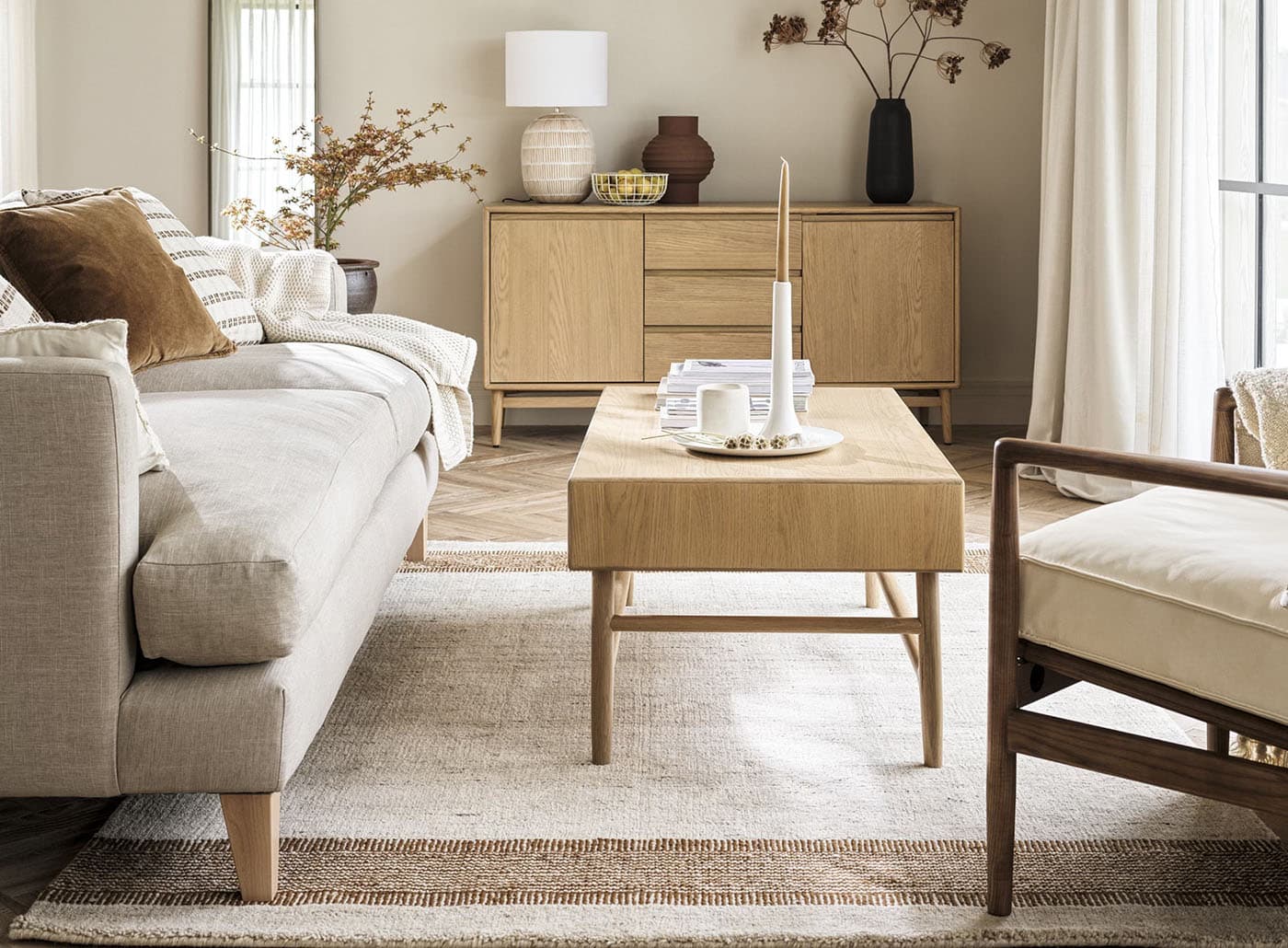 Modern living room with a beige sofa, wooden coffee table, sideboard, and neutral decor, enhanced by natural light and dried plants.