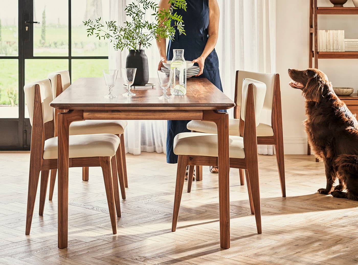 A woman sets a dining table with a plant centerpiece. Four wooden chairs surround the table, with a large dog watching her.