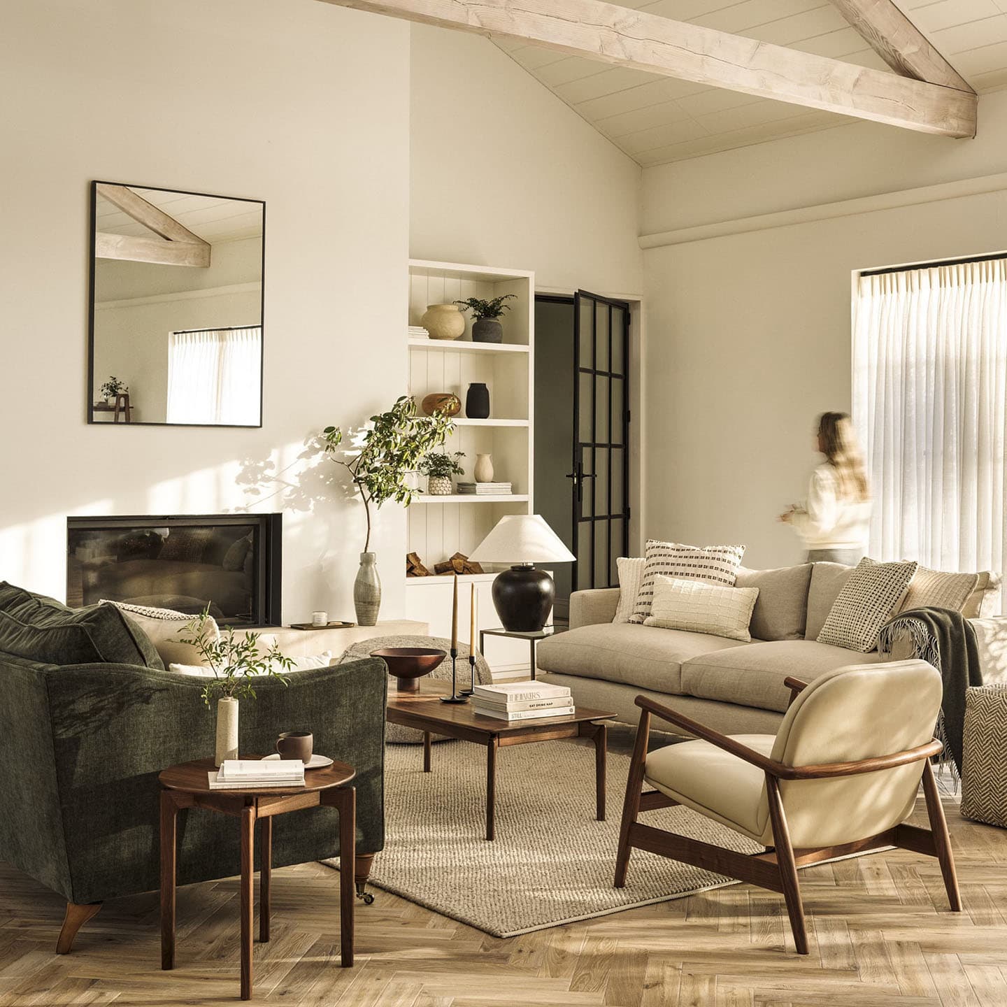 Cozy living room with neutral tones, featuring a beige sofa, armchairs, a coffee table with books, a large plant, and a person walking by.