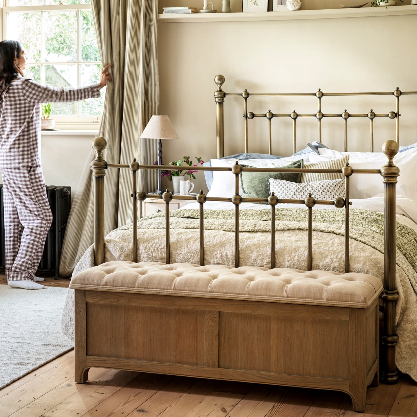 A person in pajamas opens curtains in a bedroom with a metal bed, patterned bedding, a cushioned bench, and a table lamp.