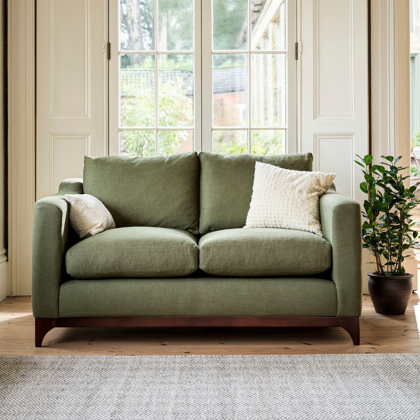 Green sofa with white cushions in front of a large window. A potted plant sits to the right on a wooden floor with a light rug.