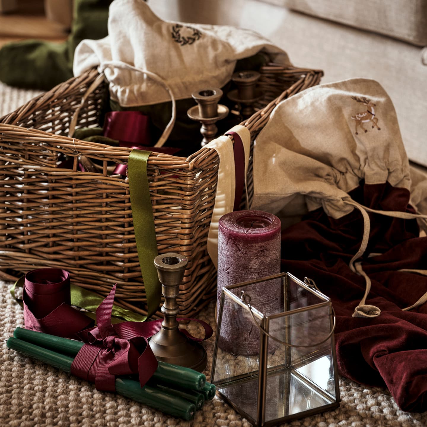 A wicker basket filled with fabric bags, brass candlesticks, green and burgundy candles, ribbons, and a glass lantern on a woven rug.