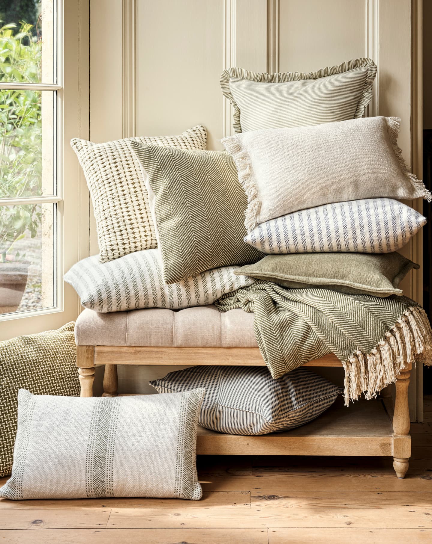 A stack of neutral-toned pillows with various textures and patterns on a wooden bench, near a sunlit window with wooden flooring.