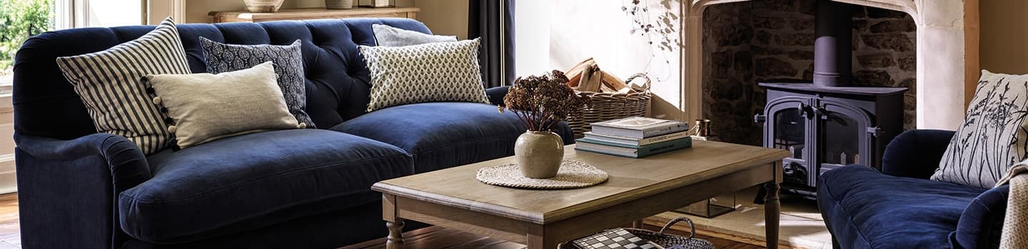 Cozy living room with a navy velvet sofa, patterned cushions, a wooden coffee table, and a stone fireplace with a wood-burning stove.