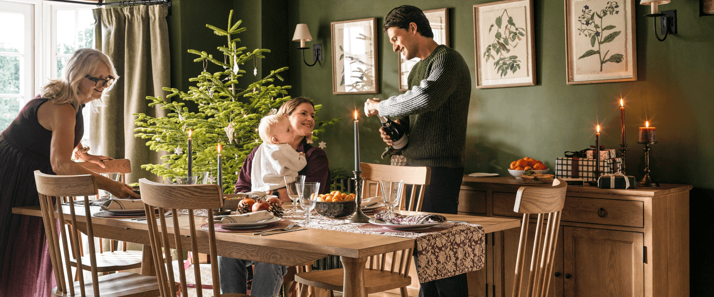 Family celebrating around a decorated dinner table with a Christmas tree, candles, and gifts in a warmly lit dining room.