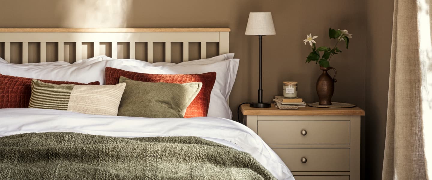 Bedroom with a painted wooden headboard, layered pillows, and a plush green blanket. A nightstand holds a lamp, books, and a vase of flowers.