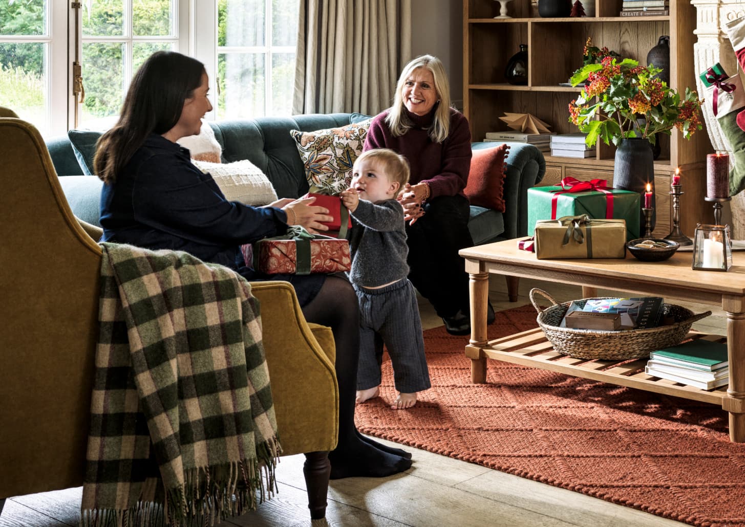 A cosy living room scene with two women and a child exchanging gifts near a decorated table, basking in natural light through large windows.