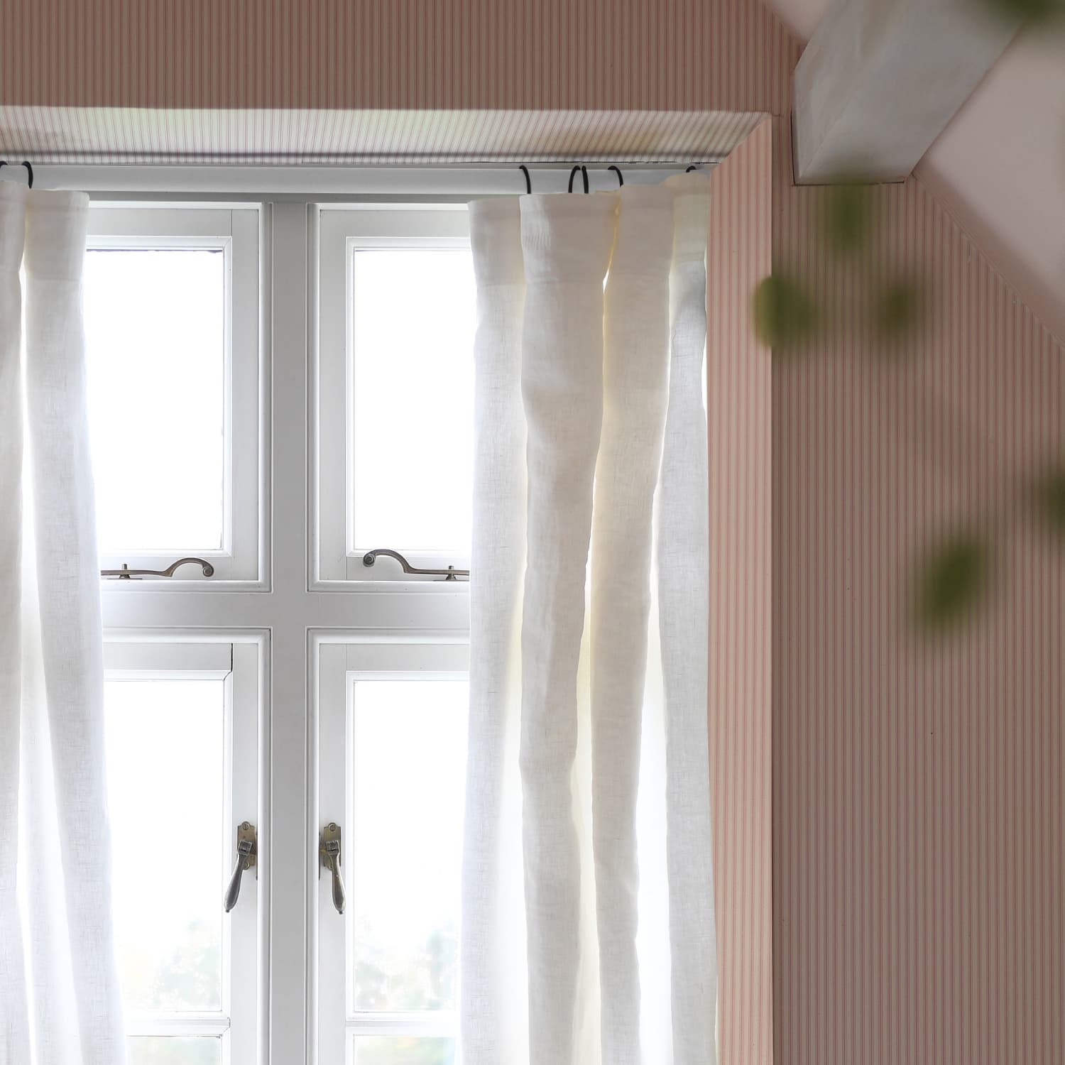 Window with white curtains partially open, allowing soft light into a room with light pink walls and a wooden beam.