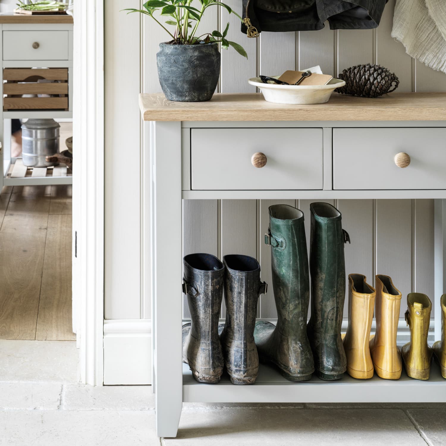 Light grey console table with two drawers and wooden top, holding a plant, dish, and pinecone, while boots are neatly arranged on the bottom shelf.
