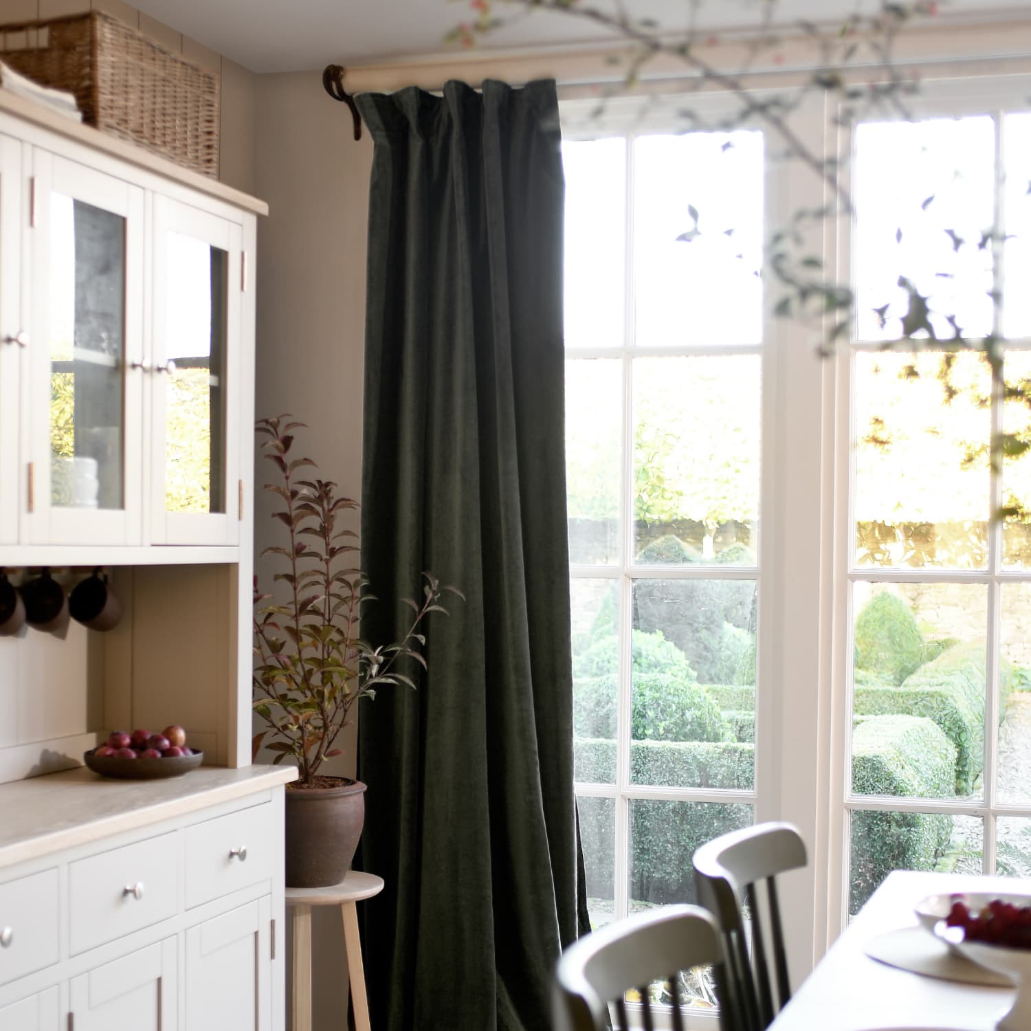 Cosy kitchen with white cabinets, a potted plant, dark curtains, and a view through large windows to a lush garden.