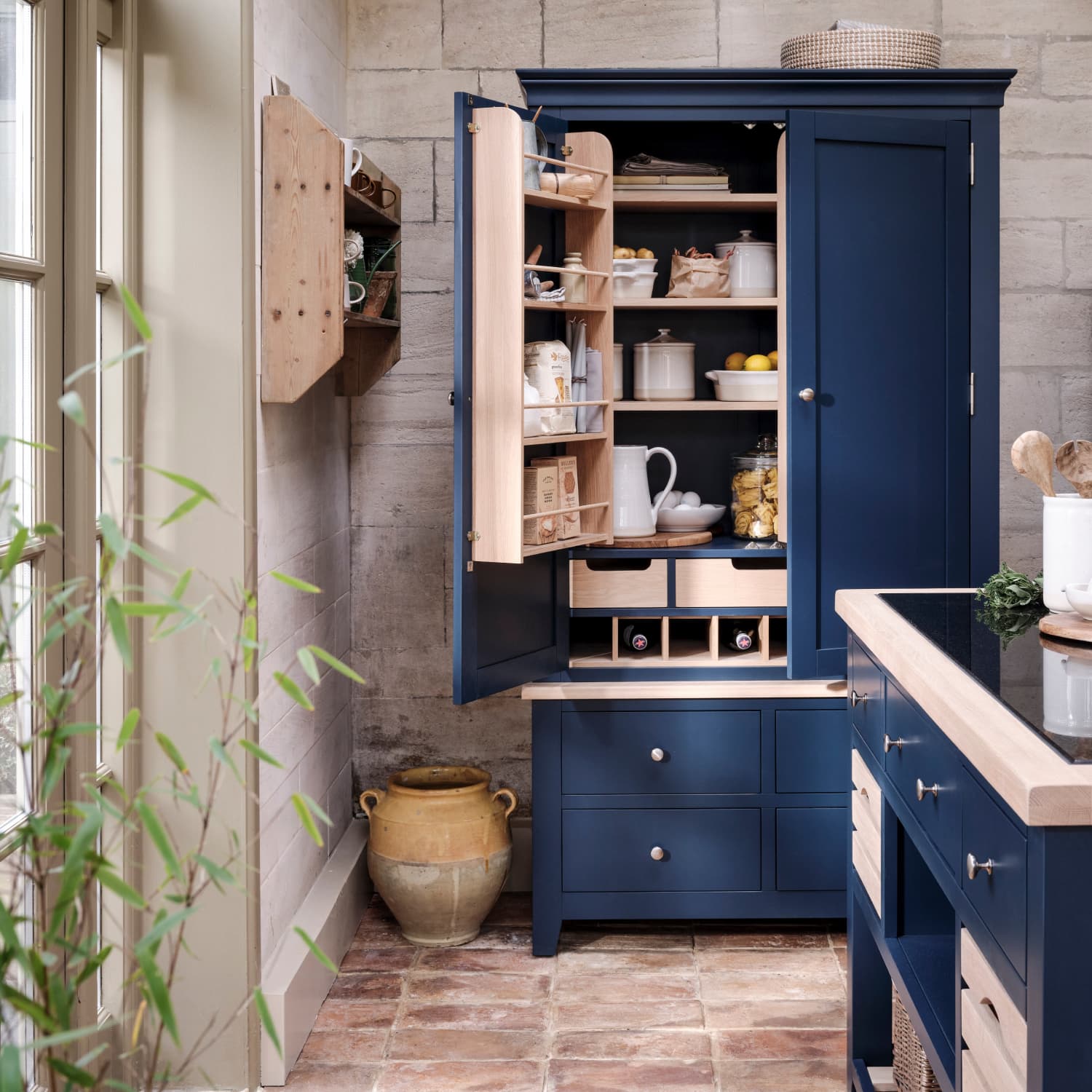 A stylish kitchen corner with a blue pantry cabinet filled with neatly arranged jars, crockery, and spices. A rustic pot sits on the brick floor.