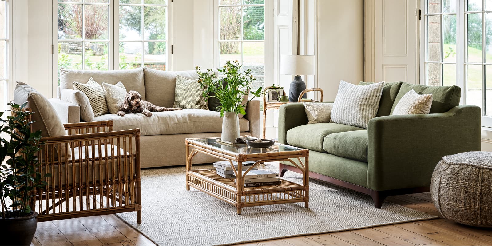 A living room with a beige sofa, green couch, rattan chair, and glass top coffee table, featuring pillows and a dog. Large windows provide natural light.