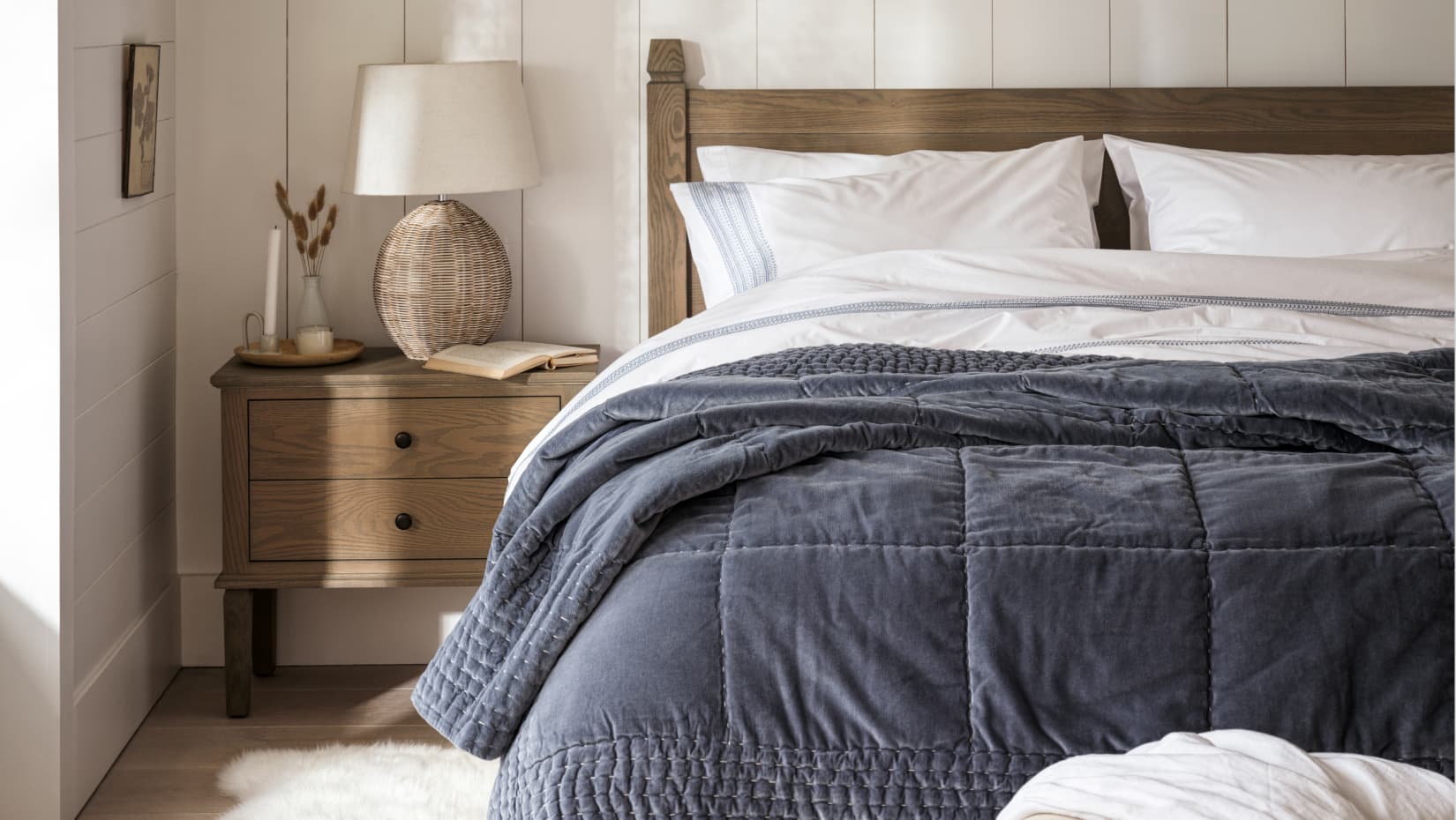 Cozy bedroom with a wooden bed, white linens, and a gray quilt. A wicker lamp and books sit on the bedside table.