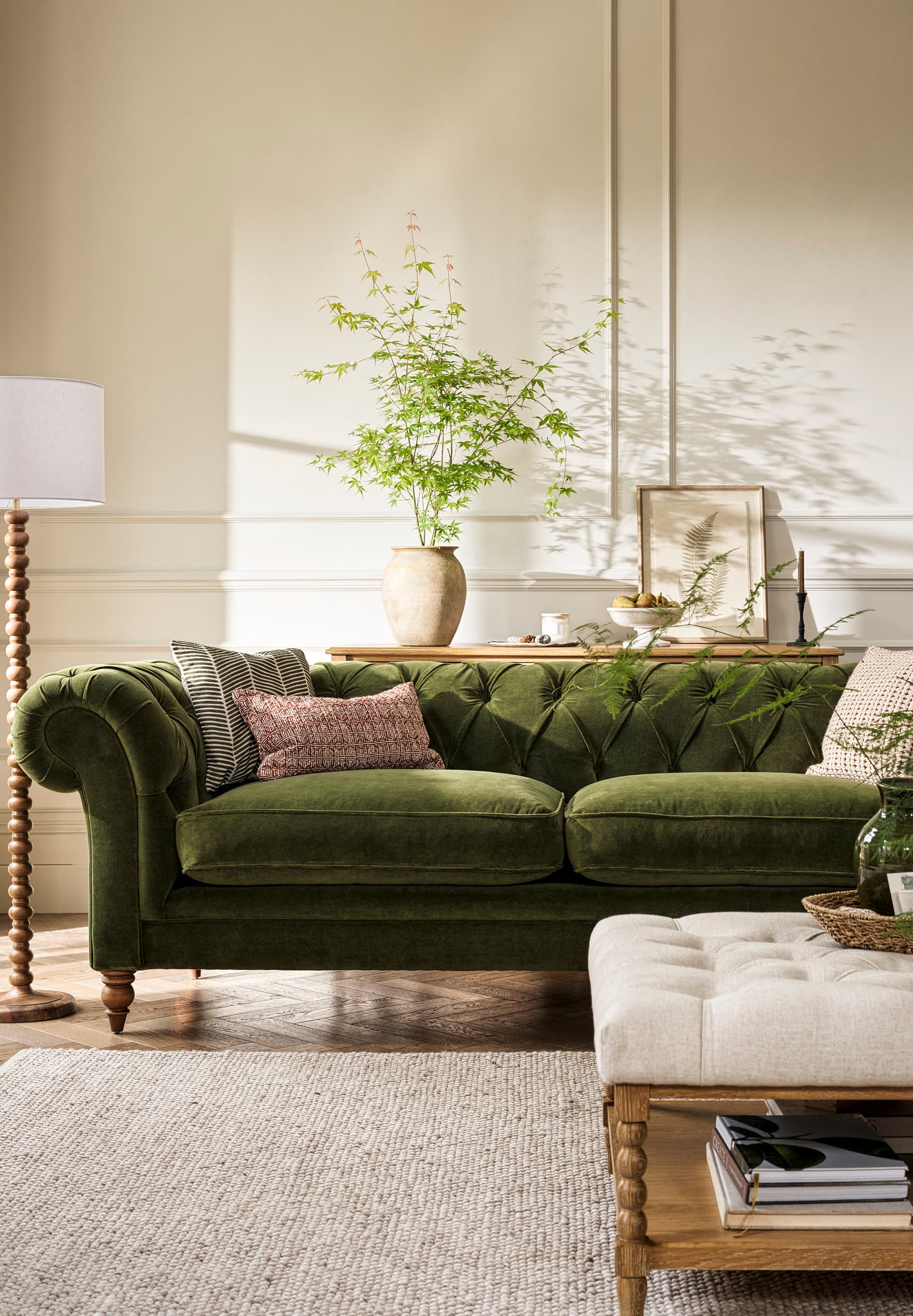 A cosy living room with a green velvet sofa, decorative pillows, and a lamp. A wooden table holds a vase with green branches and framed art.