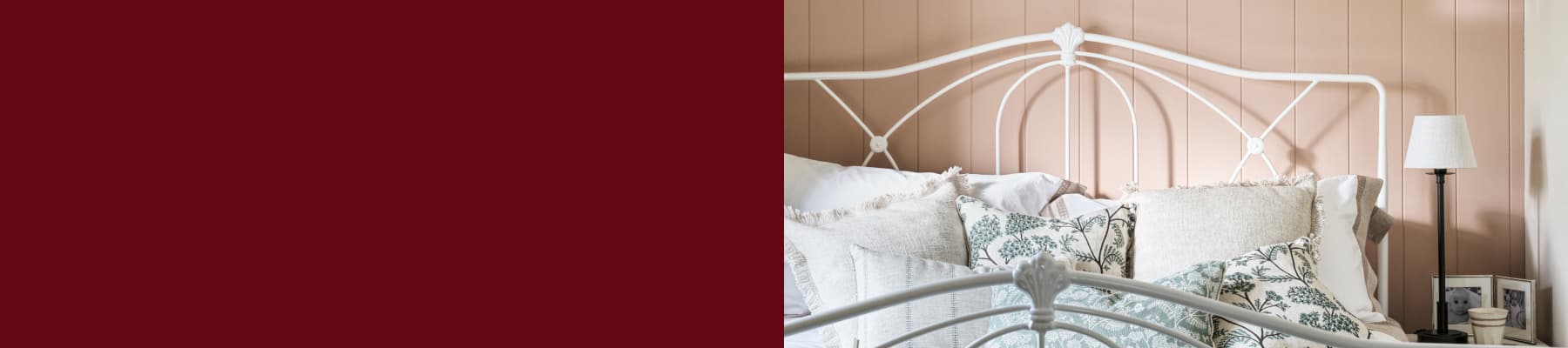 A cozy bedroom with a white metal bed, patterned and cream pillows, soft pink wall paneling, a black lamp, and framed photos on a bedside table.