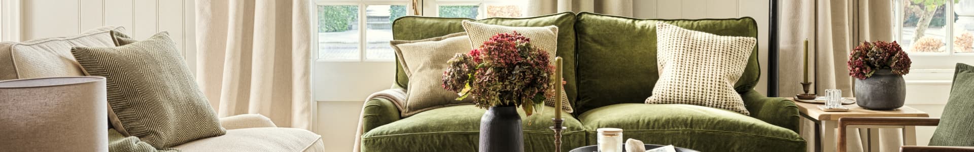 Cozy living room with a green velvet sofa, neutral pillows, dried flower arrangements, wooden tables, and beige curtains by sunlit windows.