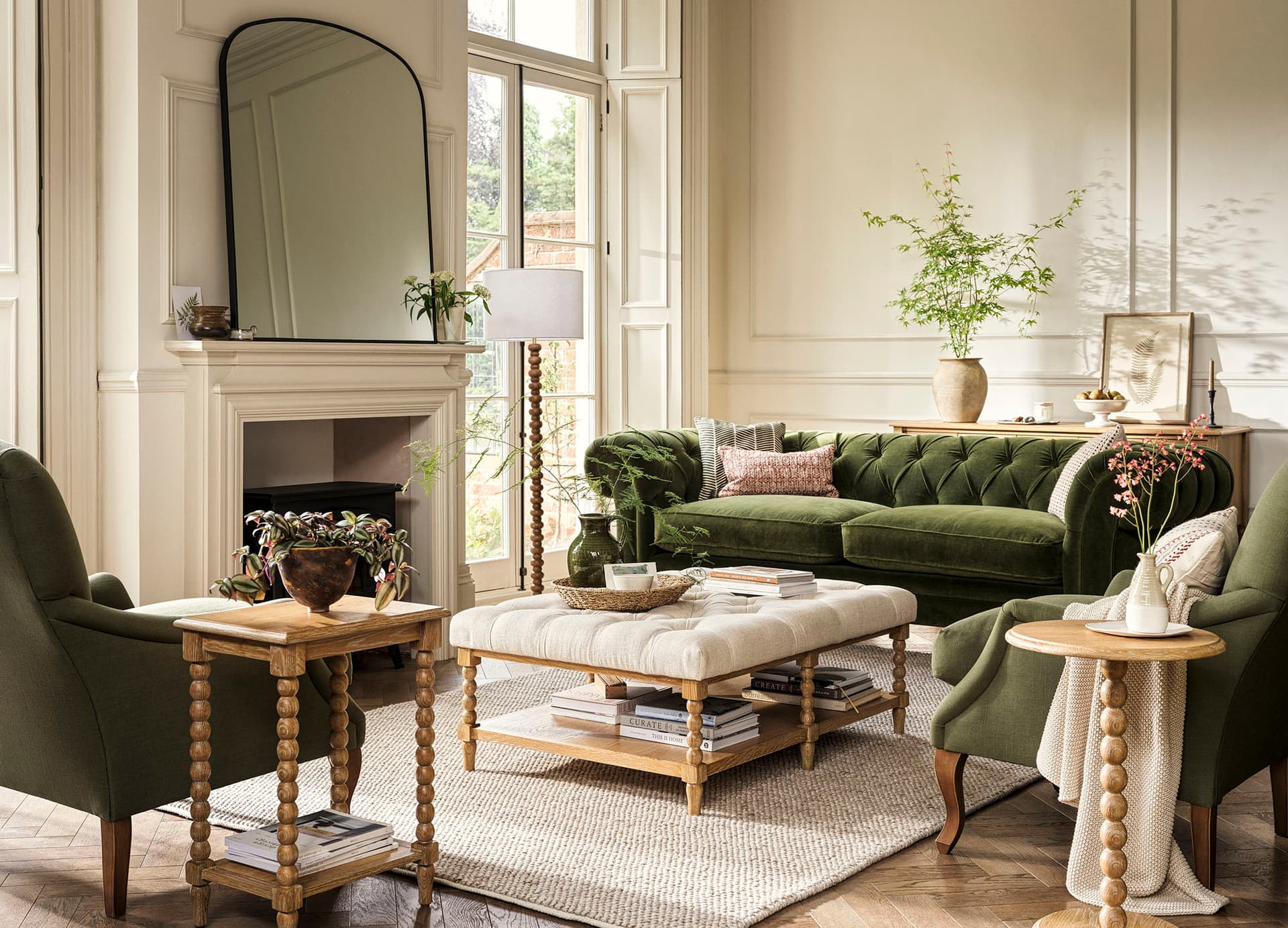 Elegant living room with green velvet sofa, armchairs, and wooden tables. A large mirror above the fireplace, surrounded by plants and decor.