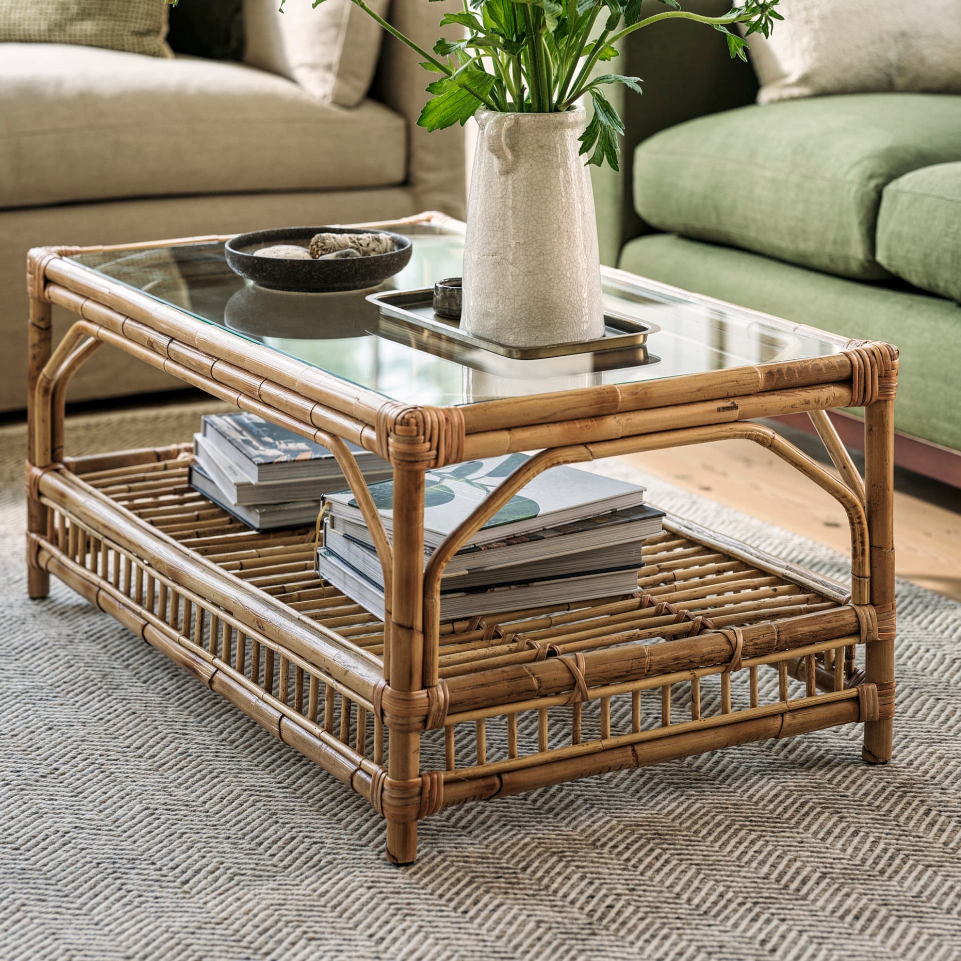 A bamboo coffee table with a glass top, books on the lower shelf, and a ceramic pitcher with greenery on top, placed on a patterned rug.