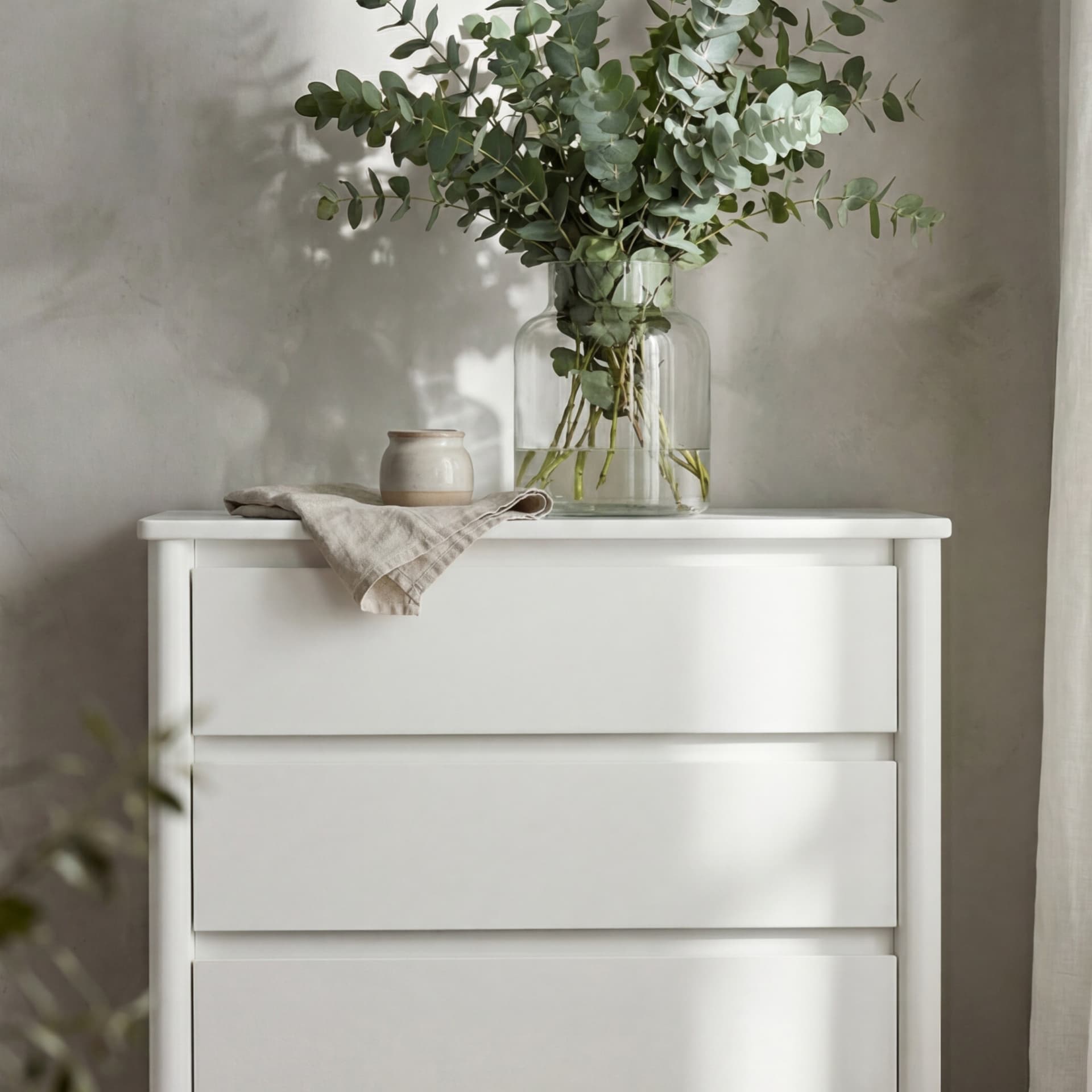 White dresser with a glass vase of eucalyptus branches, a small ceramic pot, and a folded cloth on top, against a soft light gray wall.