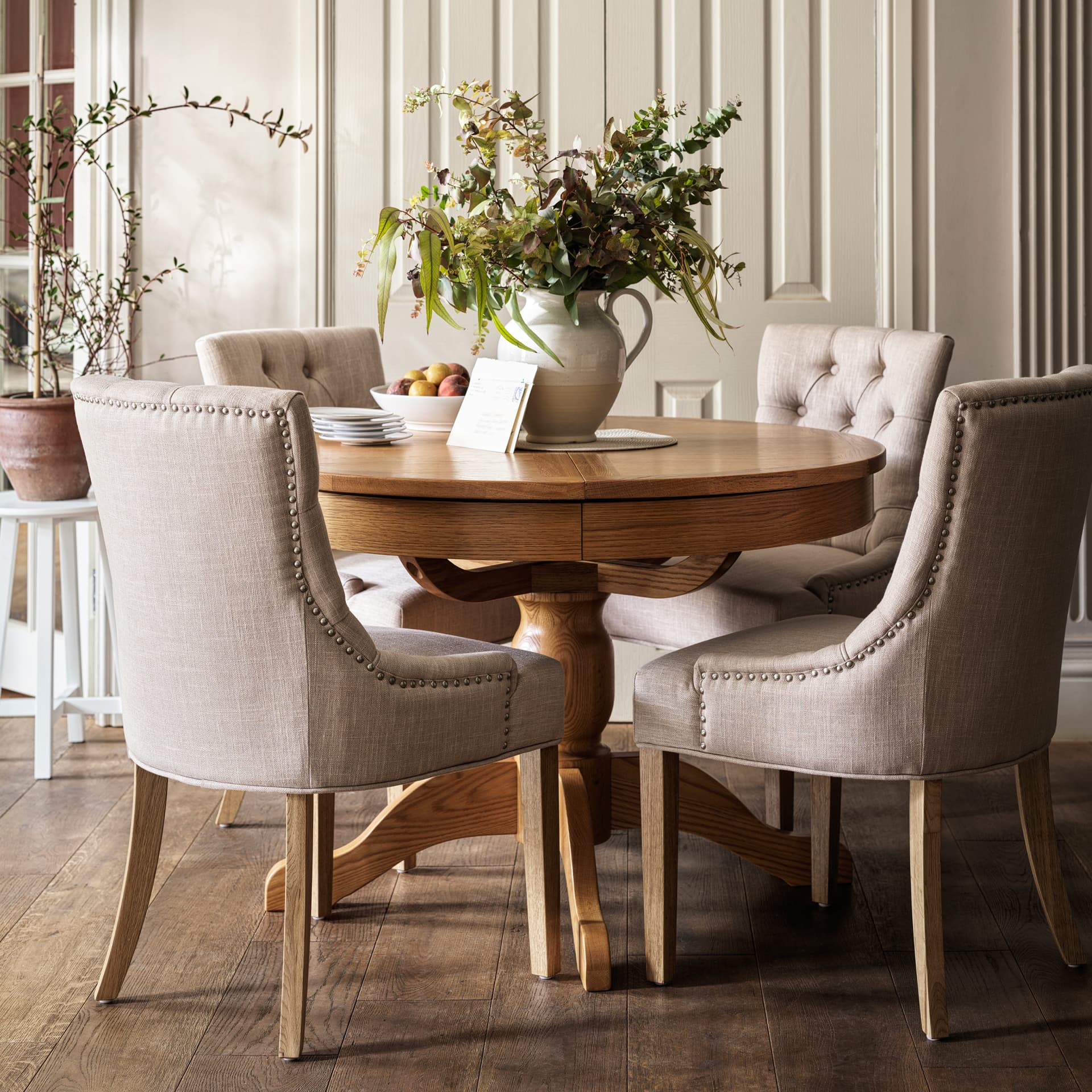 Elegant dining area with a round wooden table, beige upholstered chairs, and a vase of greenery. Hardwood floors and soft lighting enhance the cozy ambiance.