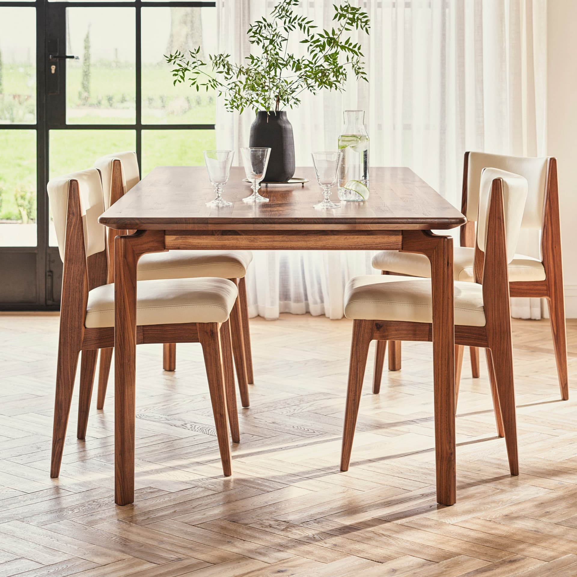 A wooden dining table set with four chairs, a black vase with green foliage, glasses, and a water carafe, placed near a large window with sheer curtains.