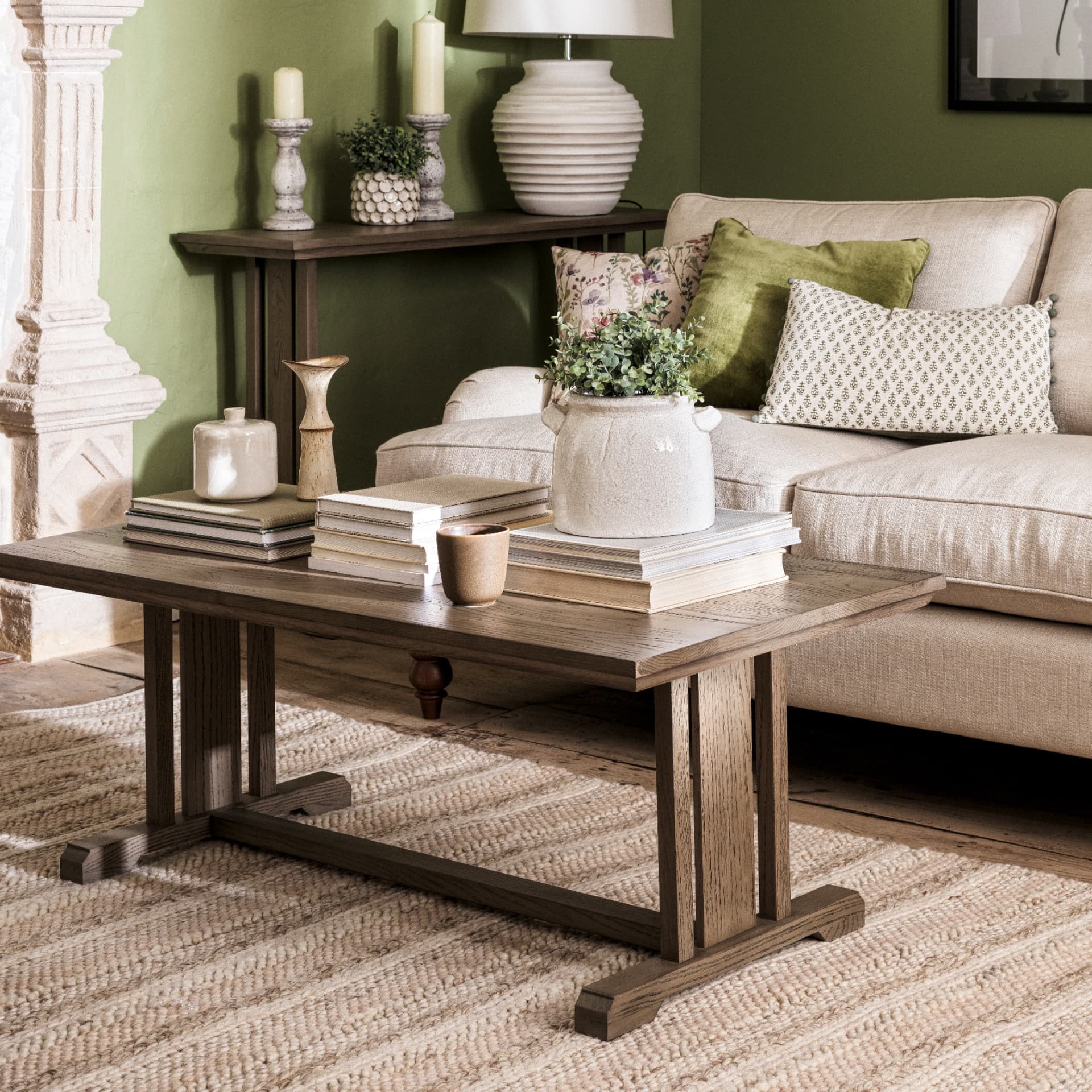 Cozy living room with a beige sofa, green pillows, wooden coffee table, books, candles, potted plant, and green wall. Warm, rustic decor.